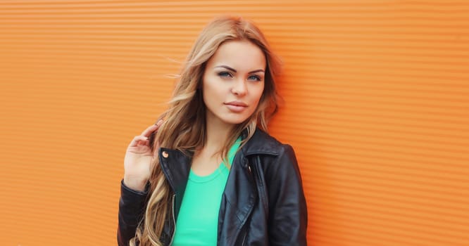 Portrait of beautiful young blonde woman posing in black leather jacket looking at camera on colorful orange background