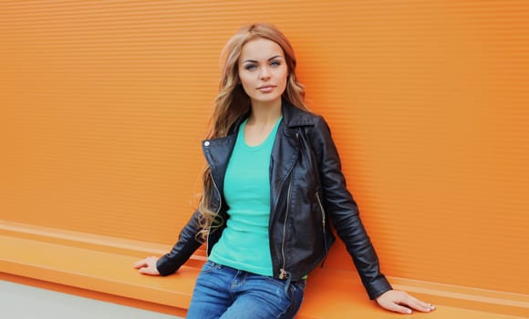 Portrait of beautiful young blonde woman posing in black leather jacket looking at camera on colorful orange background