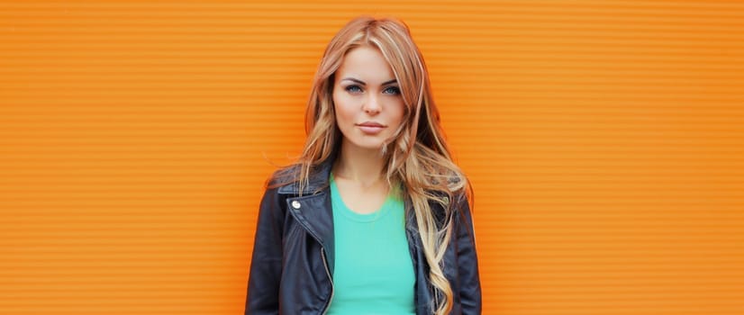 Portrait of beautiful young blonde woman posing in black leather jacket looking at camera on colorful orange background
