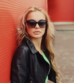 Portrait of beautiful young blonde woman posing in black leather jacket, sunglasses looking at camera on colorful red background