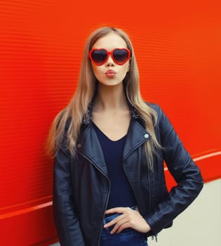 Portrait of beautiful stylish blonde young woman model blowing kiss in red heart shaped sunglasses, black rock leather jacket on city street