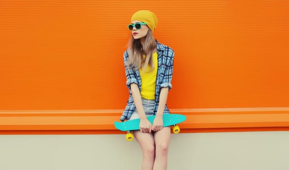 Summer portrait of stylish young woman with skateboard in colorful clothes, yellow hat looking away posing on orange background on city street