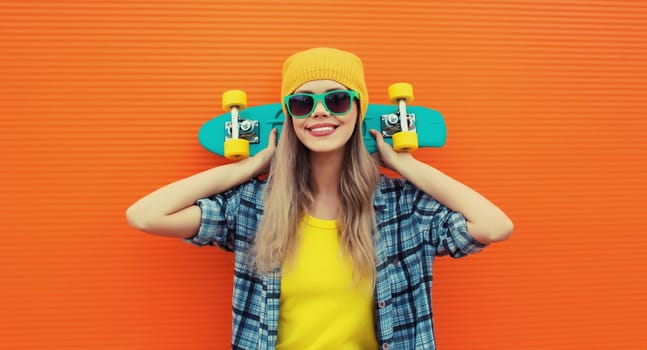 Summer portrait of happy cheerful stylish young woman with skateboard in colorful clothes, yellow hat posing on orange background on city street