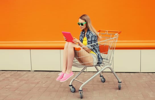 Modern stylish young woman using digital tablet computer looking at device sitting in shopping trolley cart on city street