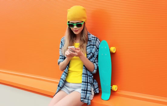 Modern stylish young woman using smartphone looking at device in colorful clothes with skateboard on city street on orange wall background
