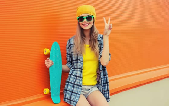 Summer portrait of happy cheerful stylish young woman with skateboard in colorful clothes, yellow hat posing on orange background on city street