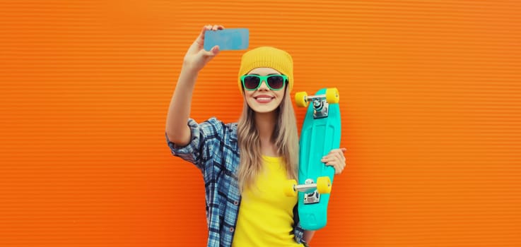 Summer portrait of happy smiling blonde young woman taking selfie with smartphone and skateboard on colorful orange background