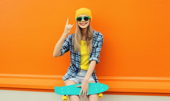 Summer portrait of happy cheerful stylish young woman with skateboard in colorful clothes, yellow hat posing on orange background on city street