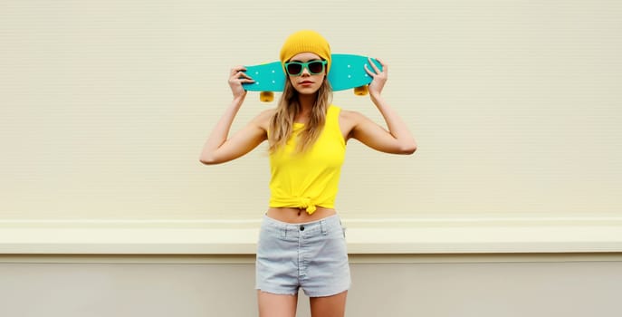 Summer portrait of stylish blonde young woman with skateboard in colorful clothes, yellow hat posing on white background on city street