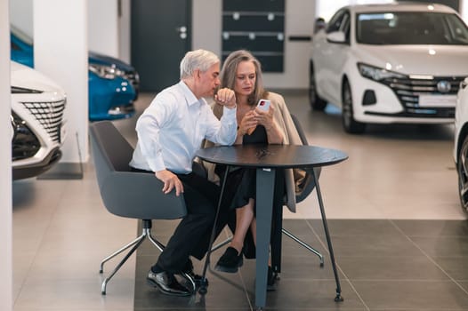 A mature couple is sitting in a car dealership and looking at a smartphone