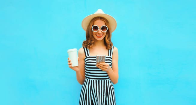 Portrait of happy smiling young woman with smartphone wearing summer straw hat, dress on blue background