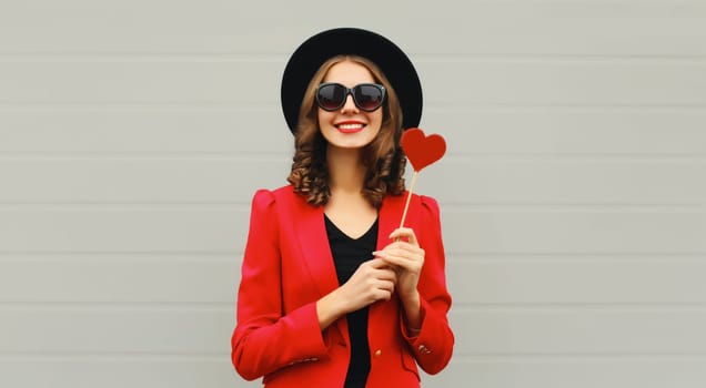 Beautiful happy woman with sweet red heart shaped lollipop on stick in black round hat