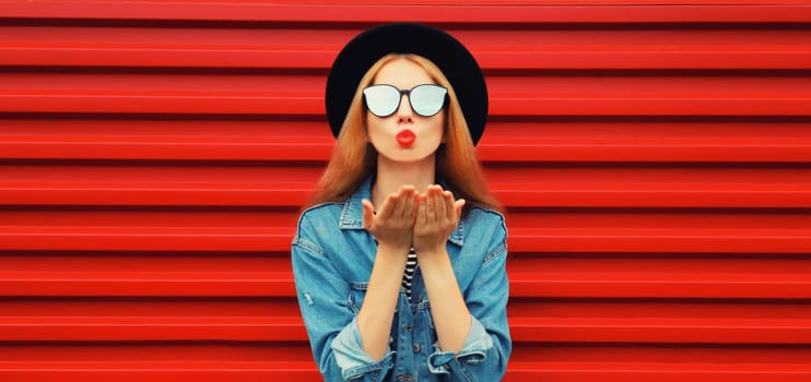 Portrait of stylish young woman model blowing her lips sending sweet air kiss wearing jean jacket, black round hat on red background