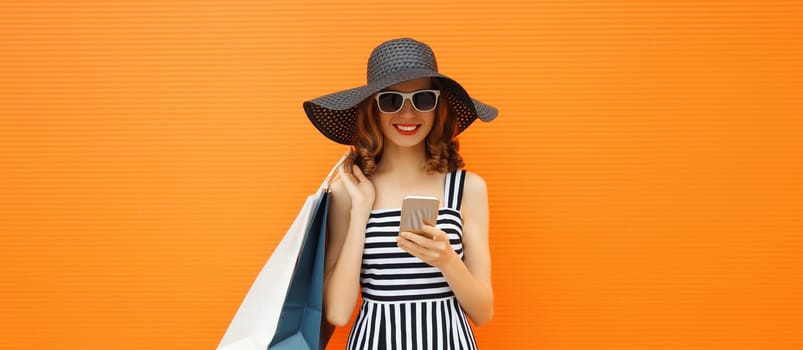 Beautiful happy young woman looking at phone with shopping bag in summer black straw hat, striped dress on orange background