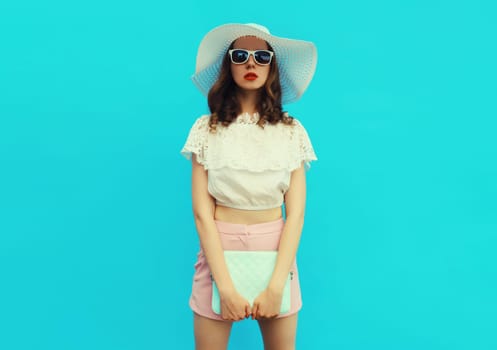 Beautiful caucasian young woman model posing with handbag clutch wearing white summer straw hat on blue studio background
