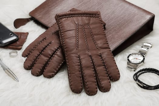 Pair of men's brown leather gloves and other men's accessories over white fur.