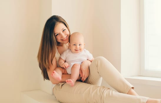 Happy smiling young mother playing with cute baby sitting together in white room at home