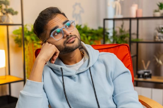 Portrait of sad Indian man guy sitting at home looks pensive thinks over life concerns or unrequited love, suffers from unfair situation. Problem, break up, depressed feeling bad annoyed, burnout