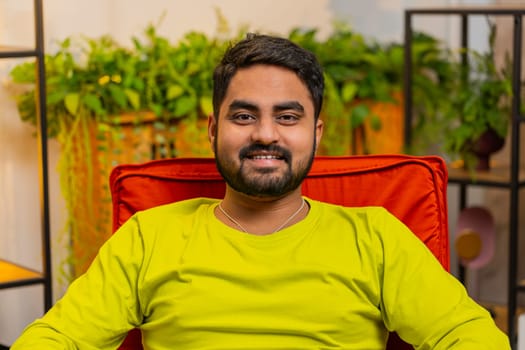 Portrait of happy calm Indian man at home chair smiling friendly, glad expression looking away dreaming resting, relaxation feel satisfied good news, celebrate win. Arabian young guy in living room