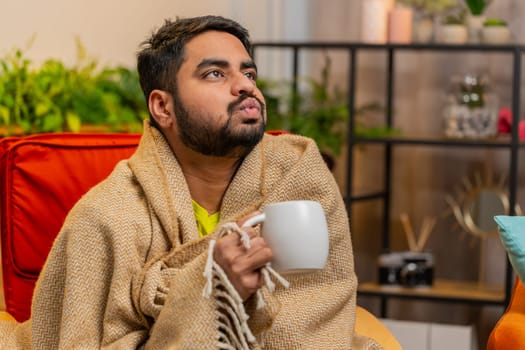 Sick Indian man in plaid sit alone shivering from cold on sofa drinking hot tea in unheated apartment without heating due debt. Unhealthy Arabian guy sits on chair feeling discomfort try to warming up