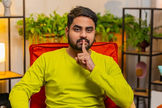 Shh be quiet please. Indian Hindu confident man presses index finger to lips makes silence hush gesture sign do not tells gossip secret. Arabian guy at home apartment living room sitting on chair