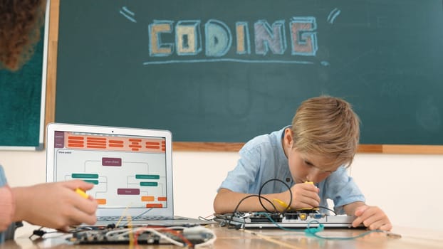 Caucasian smart boy fixing main board by using screwdriver at stem technology lesson while smiling student working together to inspect electronic machine at table with laptop display code. Pedagogy.