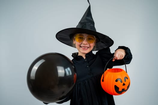 Portrait of a little Caucasian girl in a witch costume holding a cauldron and a black balloon on a white background