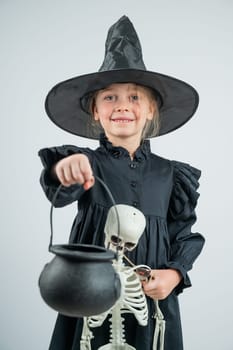 Portrait of a little Caucasian girl in a witch costume holding a cauldron and a skeleton on a white background. Vertical photo