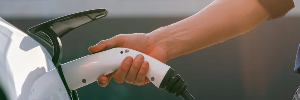 Young man put EV charger to recharge electric car's battery from charging station in city commercial parking lot with sunlight. Rechargeable EV car for sustainable urban travel. Panorama Expedient