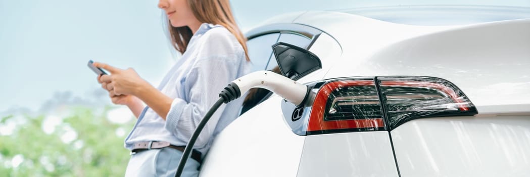 Young woman using smartphone online banking application to pay for electric car battery charging from EV charging station during vacation road trip at national park or summer forest. Panorama Exalt