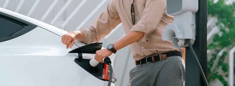 Young man recharge electric car's battery from charging station in city commercial parking lot. Rechargeable EV car for sustainable environmental friendly urban travel. Panorama Expedient