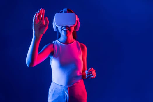 Smart female standing with surrounded by cyberpunk neon light wear VR headset connecting metaverse, futuristic cyberspace community technology. Woman using hand touching virtual object. Hallucination.