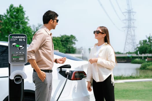 Young couple recharge EV car battery at charging station connected to power grid tower electrical industrial facility as electrical industry for eco friendly vehicle utilization. Expedient