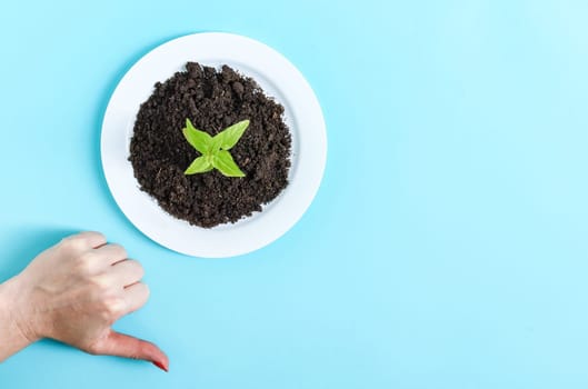 A plastic white plate with black loose soil, a green sprout and a female caucasian hand with a dislike sign on the left on a blue background with copy space on the right, flat lay close-up. Concept ecology and plastic waste.