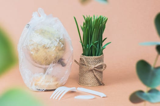 One planet earth globe inside a transparent plastic bag with a flower in a pot, a broken disposable plastic fork and eucalyptus branches on a beige background, side view close-up.