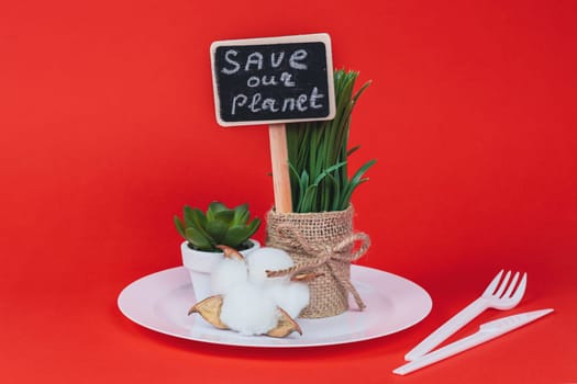 One baggy pot with sprouted wheat, a succulent on a white plastic plate with a peg with the inscription: save our planet and a disposable fork and knife stand in the center on a red background, side view close-up.