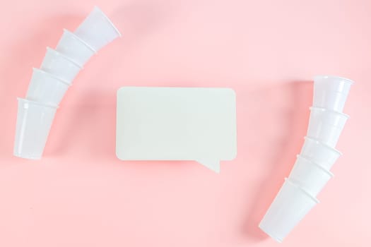 Ten disposable white glasses and an empty callout lie on a pastel pink background with space for your text, flat lay close-up.