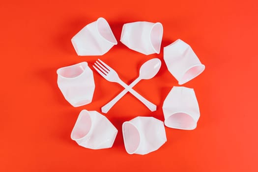 Six plastic crumpled glasses are laid out in the center in a circle with one disposable fork and spoon arranged crosswise lie on a red background as a symbol of a prohibition sign, flat lay closeup. Eco-friendly concept, disposable cutlery and ecology day.