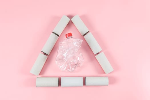 Seven empty toilet paper rolls arranged in a triangle with a crumpled plastic transparent bottle on a pastel pink background close up, flat lay closeup. Ecology concept, waste sorting and recycling.
