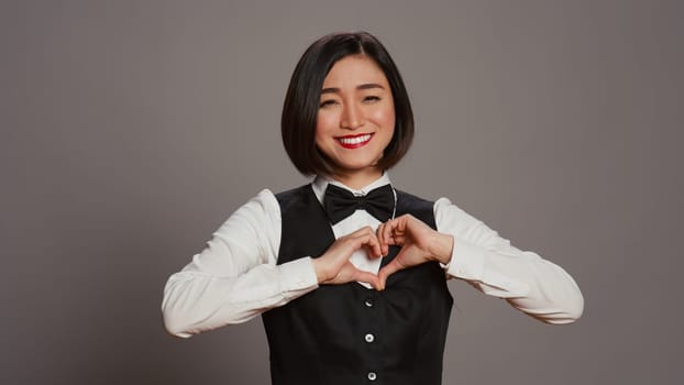 Hotel administrator shows heart shape symbol on camera, expressing romantic gesture and being flirty. Asian receptionist presenting true honest feelings, does romance sign in studio. Camera B.