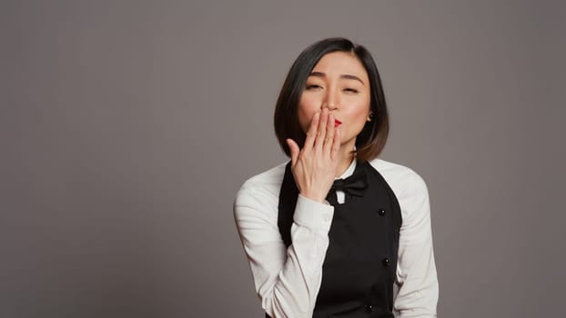 Cute flirty waitress blowing air kisses in front of camera, acting romantic with sweet gesture. Asian restaurant employee posing with a kissy facial expression, catering industry. Camera B.