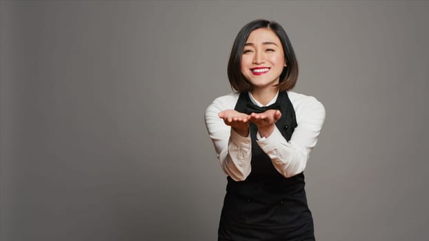 Cute flirty waitress blowing air kisses in front of camera, acting romantic with sweet gesture. Asian restaurant employee posing with a kissy facial expression, catering industry. Camera A.