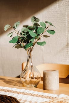 One glass transparent vase with eucalyptus branches and a candle stand on a wooden kitchen table against the wall on a summer evening, close-up side view.