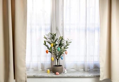 Easter marble decorative eggs hang on the branches of a green tree in a pot that stands on the sill of a window in the living room early in the morning, close-up side view.