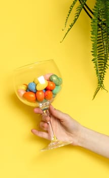 The hand of a young Caucasian unrecognizable girl holds a wine glass with Easter eggs and marble decorative eggs on a yellow background with a hanging palm flower, close-up side view.