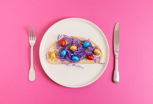One sandwich with decorative lilac paper hay with chocolate Easter eggs in shiny multi-colored wrappers on a tray with a fork and knife lie in the center on a pink background, flat lay close-up. Creative Easter food concept.