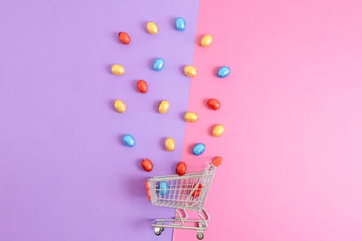 Chocolate Easter eggs in shiny multi-colored wrappers spill out of a mini shopping cart, lie in the center on a lilac-pink background with copy space on the sides, flat lay close-up. Easter shopping concept.