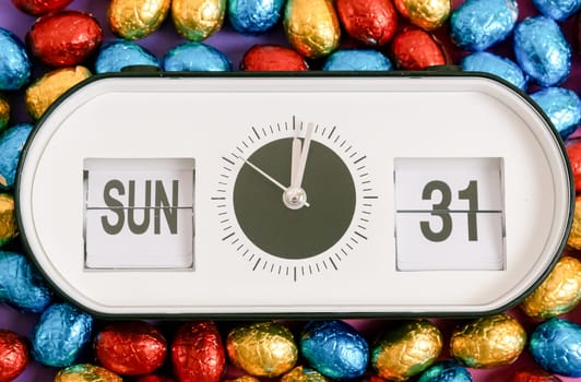 One alarm clock with date, time of Easter 2024: Sunday March 31st with chocolate Easter eggs in a colorful shiny wrapper on a lilac-pink background, flat lay very close-up. Concept of Easter candy, holiday time.