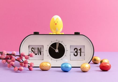One alarm clock with date, time of Easter 2024: Sunday March 31st with chocolate colorful Easter eggs, Easter chick and pussy willow branches on a lilac pink background, close-up side view. Easter candy concept, holiday time.