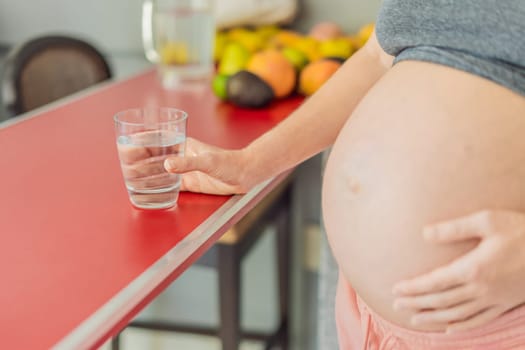 Embracing the vital benefits of water during pregnancy, a pregnant woman stands in the kitchen with a glass, highlighting hydration's crucial role in maternal well-being.